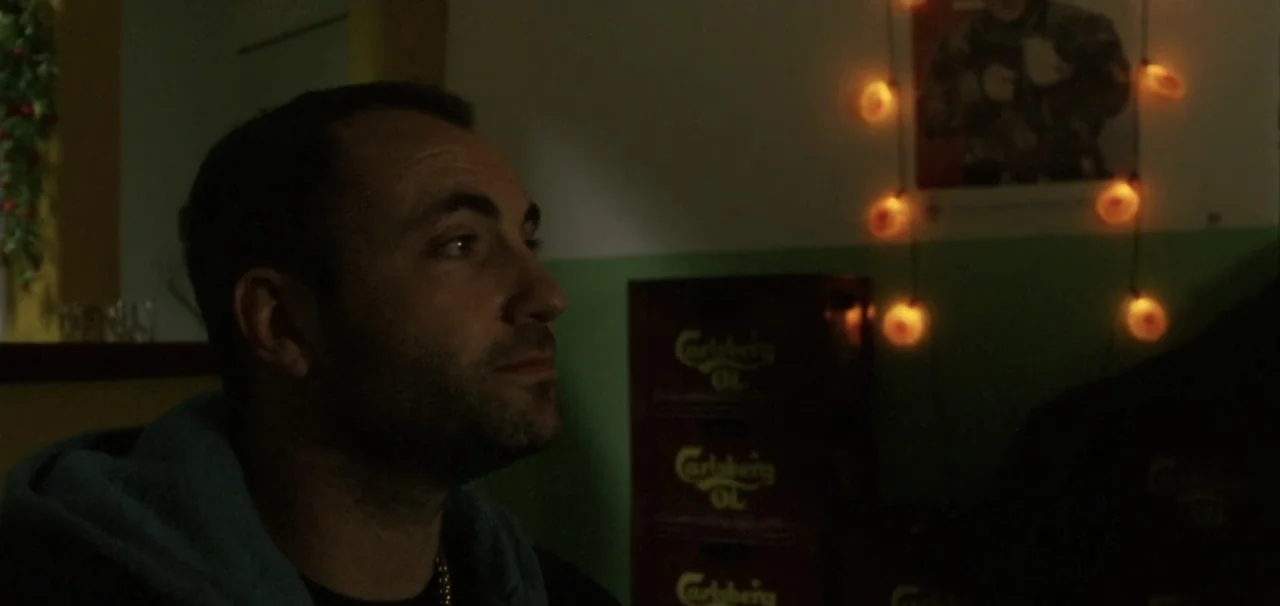 Pusher (1996) movie still: closeup — A dimly lit interior space features green walls decorated with string lights and beer crates. A man with a dark…