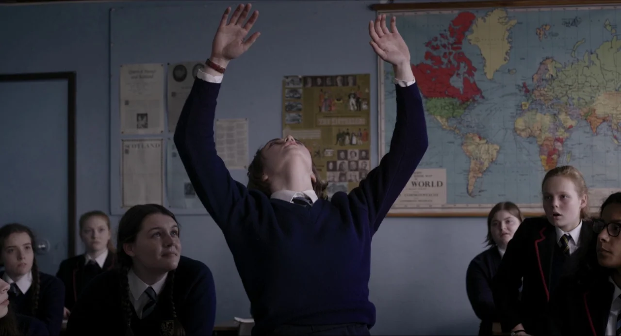 The Falling movie still: medium — A student in a school uniform sits at a desk in a classroom. The student reaches both arms towards the ceiling…