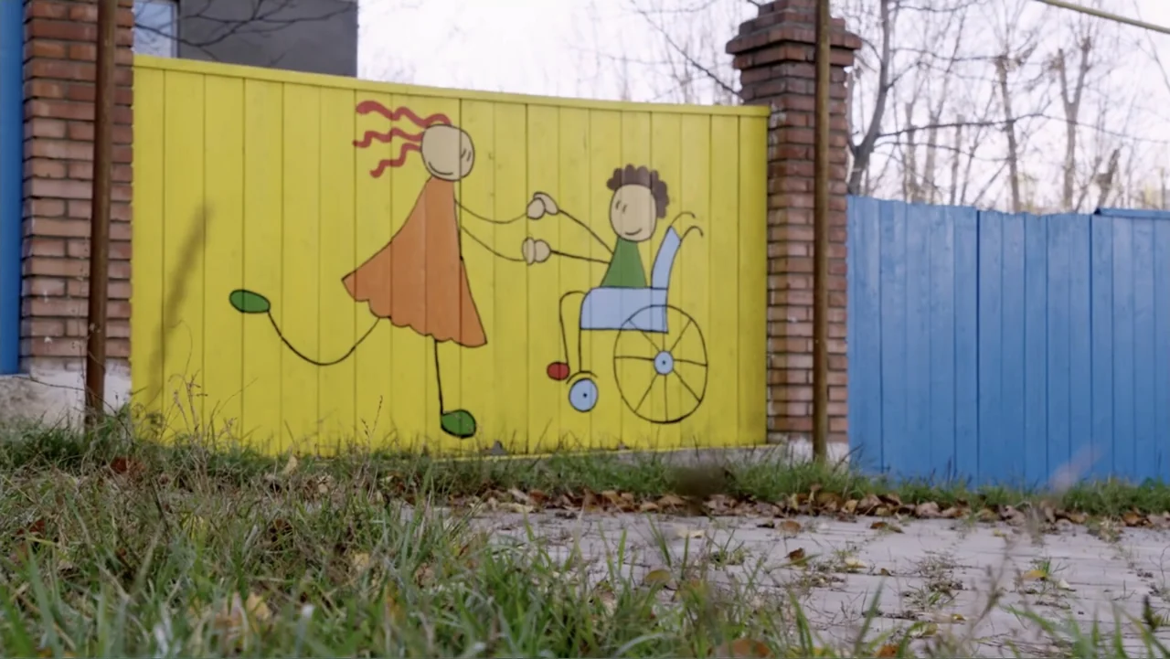 Finding the Way Home (1991) movie still: medium — A bright yellow wooden fence stands outdoors with a painted mural showing two figures. A girl…