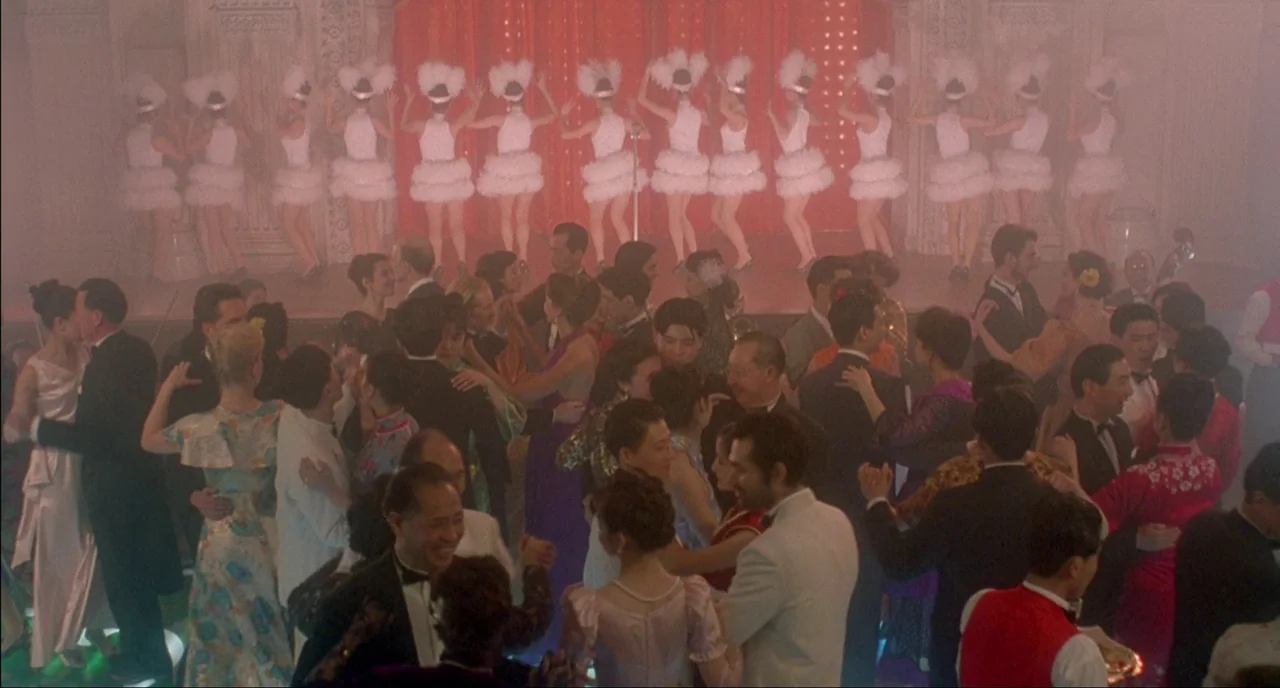 Shanghai Triad (1995) movie still: wide — A crowded 1940s ballroom with couples dancing under hazy light and red curtains. Performers in white…