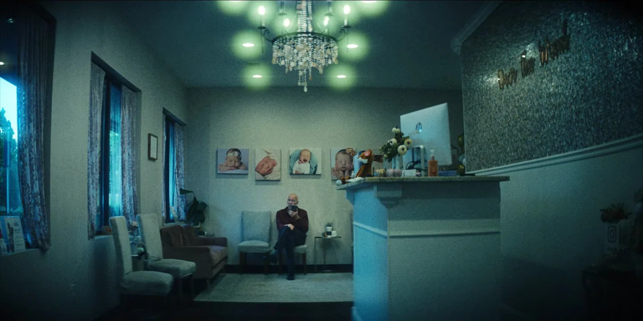 Spermworld (2024) movie still: wide — A waiting room features gray walls decorated with four framed photos of newborn babies. An older man sits…