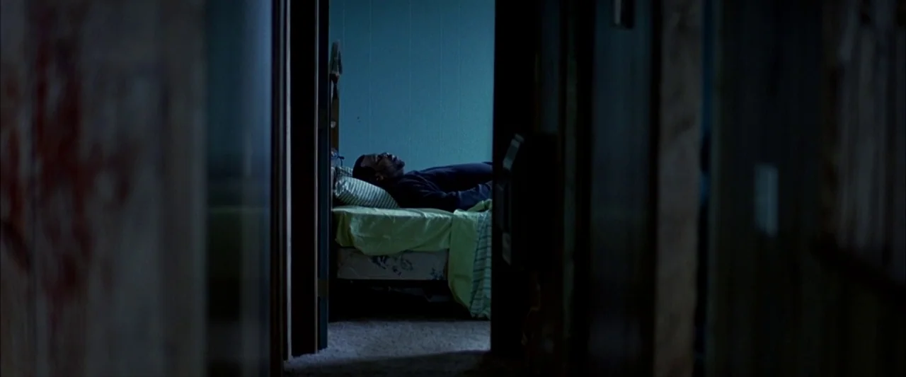 Ballast (2008) movie still: wide — A dimly lit hallway leads into a sparsely furnished room with blue walls. A man lies on a single mattress on the…