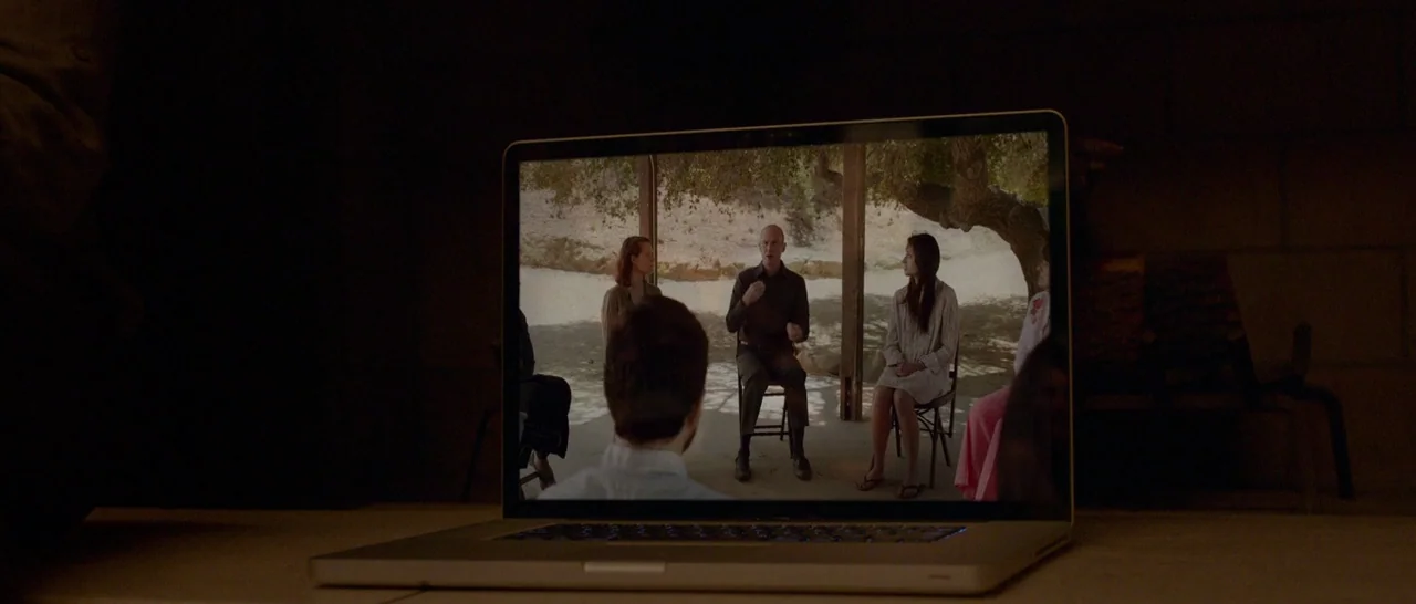 The Invitation (2015) movie still: medium — A dark, dimly lit room contains a laptop sitting on a flat surface. The screen displays a video of a…