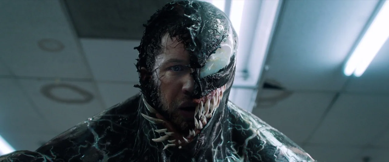Venom (2018) movie still: closeup — An interior space features ceiling tiles illuminated by cool, artificial fluorescent overhead lights. A man with…