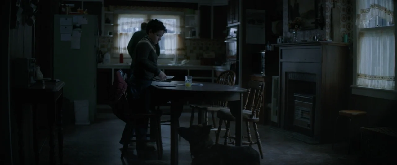 Wolf Man (2025) movie still: medium — A dimly lit room with patterned wallpaper, dark wood furniture, and two windows. A child holds a plate at a…