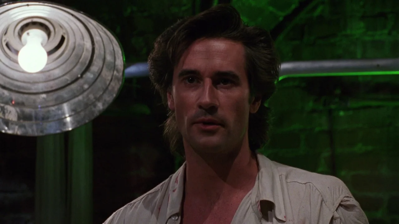 Bride of Reanimator (1989) movie still: A man stands in a dark, industrial room illuminated by a single harsh lamp. The high-contrast green lighting…