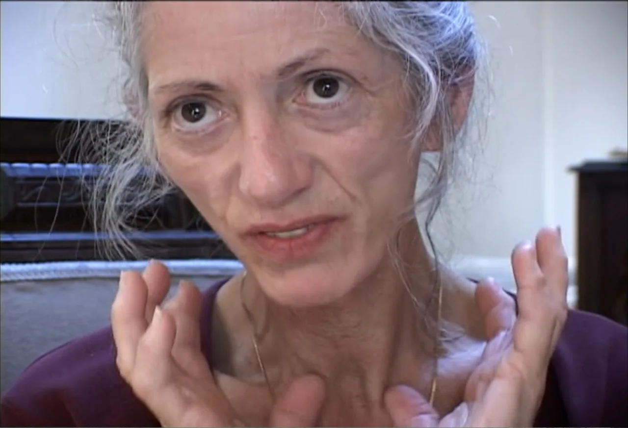 Plastic Disasters (2006) movie still: closeup — A plain interior room with a dark wooden object partially visible in the background. An elderly…