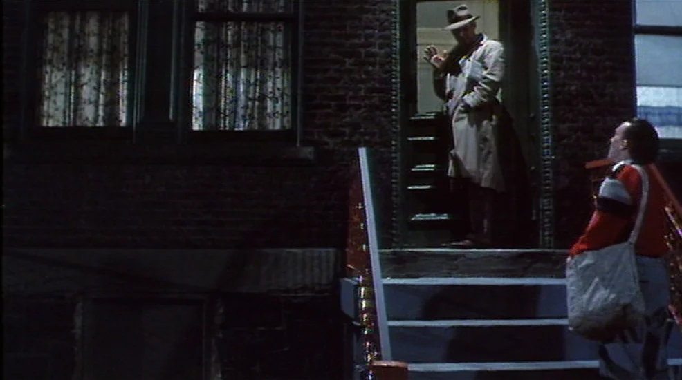 Union City (1980) movie still: wide — A brick apartment building exterior at night with an open door at the top. Two men converse while one stands…