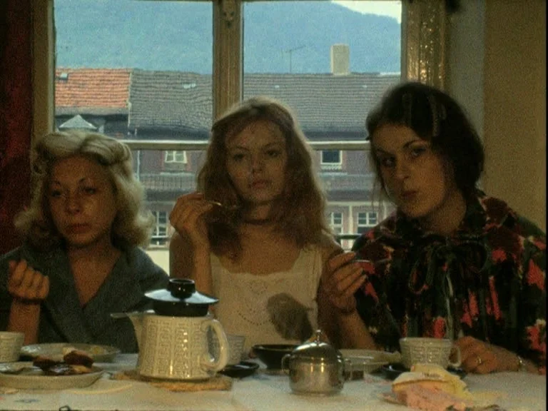 Der Bomberpilot (1970) movie still: medium — Three women sit at a dining table before a window overlooking a European cityscape. They are eating…