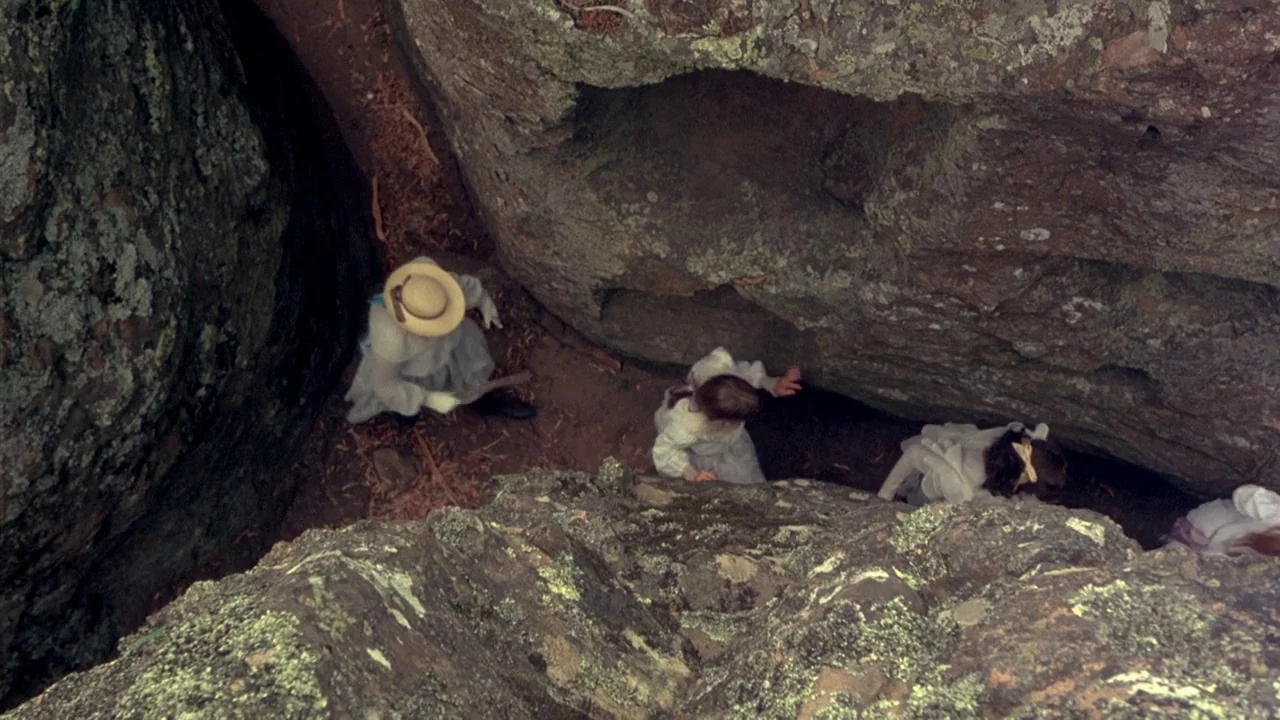 Picnic at Hanging Rock (1975) movie still: wide — Four figures dressed in long white Edwardian dresses navigate a narrow rocky crevice. They descend…