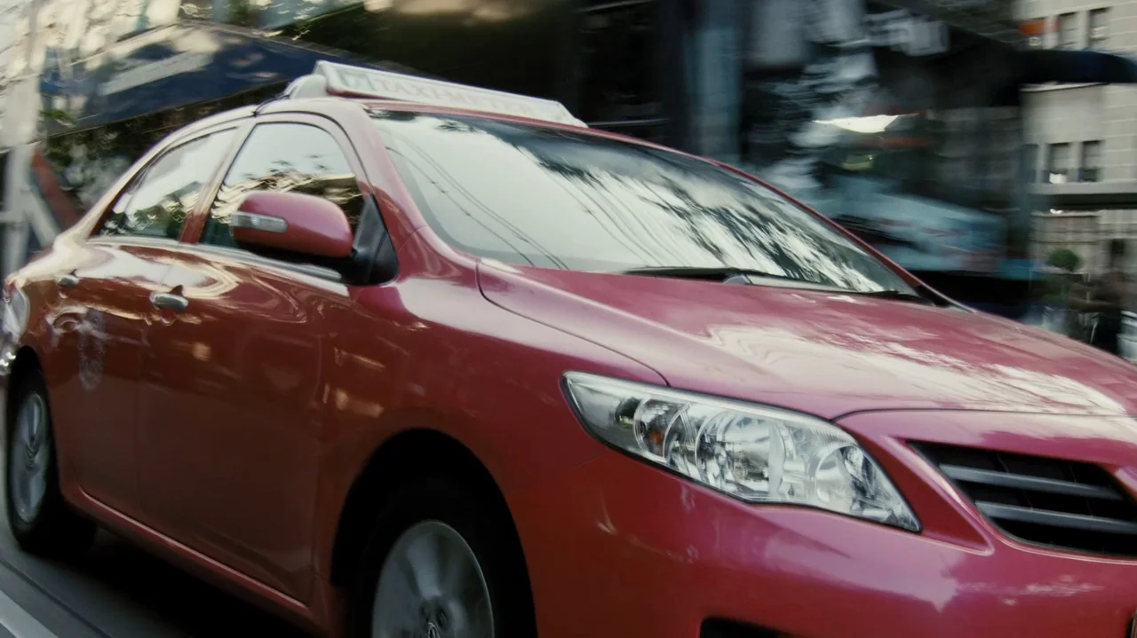 Zero Tolerance (2015) movie still: medium — A pink sedan moves rapidly along a city street lined with concrete buildings. Motion blur in the…