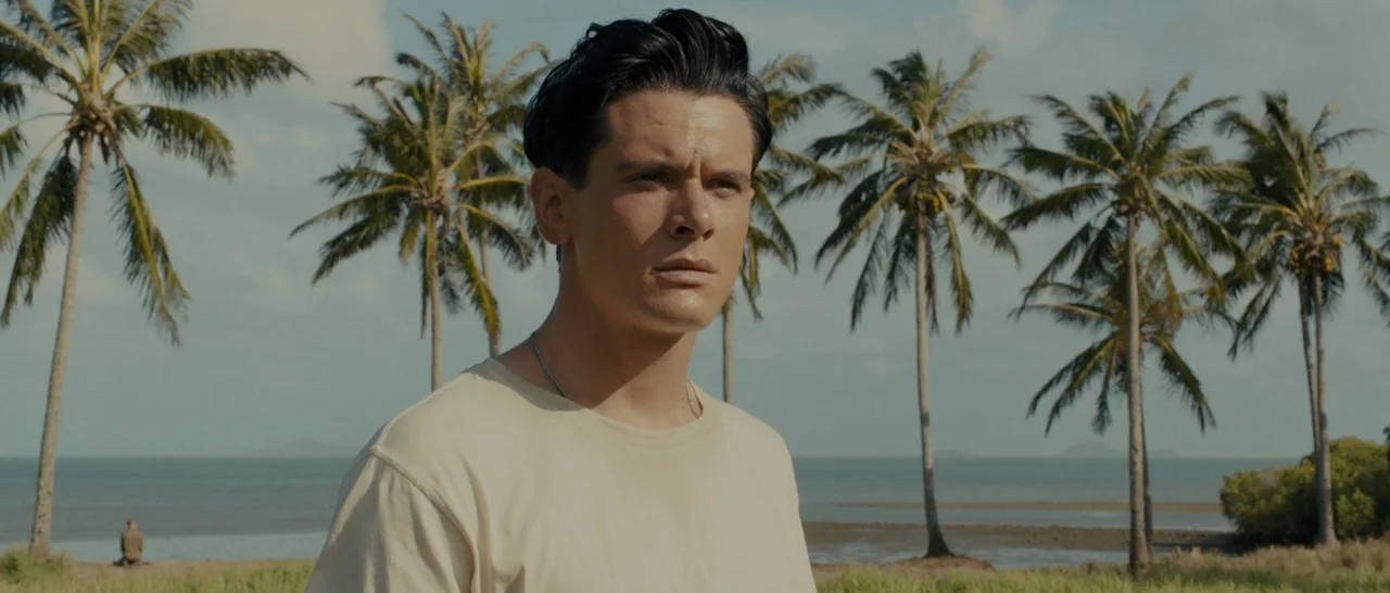 Unbroken (2014) movie still: medium — A coastal landscape features a line of tall palm trees against a calm sea. A man in a cream shirt looks toward…