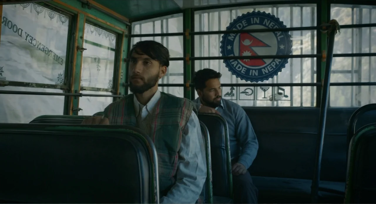 Ice Road: Vengeance (2025) movie still: medium — The interior of a bus with vinyl bench seats and a Nepal sticker visible. Two men sit on the seats…