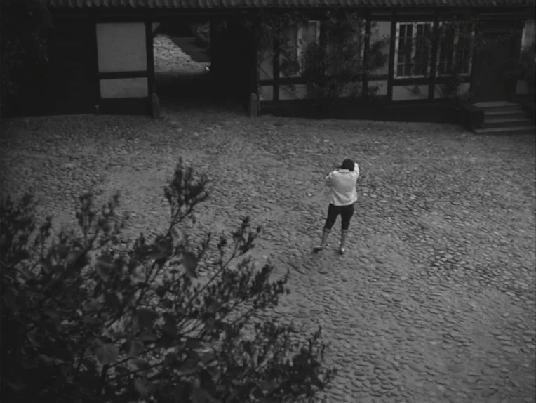 A Lesson In Love (1954) movie still: wide — A stone-paved courtyard surrounded by timber-framed structures with dark wooden beams. A person stands…