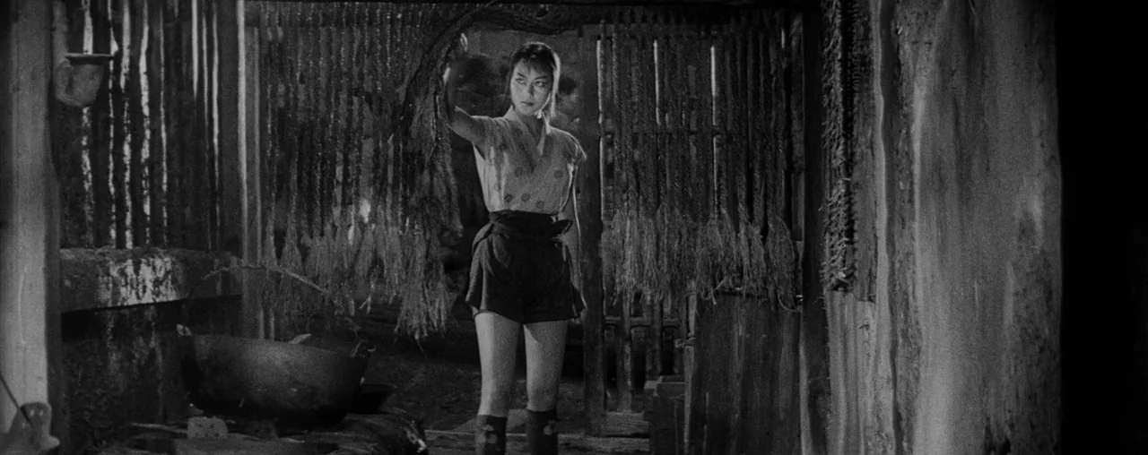 The Hidden Fortress (1958) movie still: fullbody — A rustic interior with dark wooden walls and hanging bundles of rope or straw. A young woman…