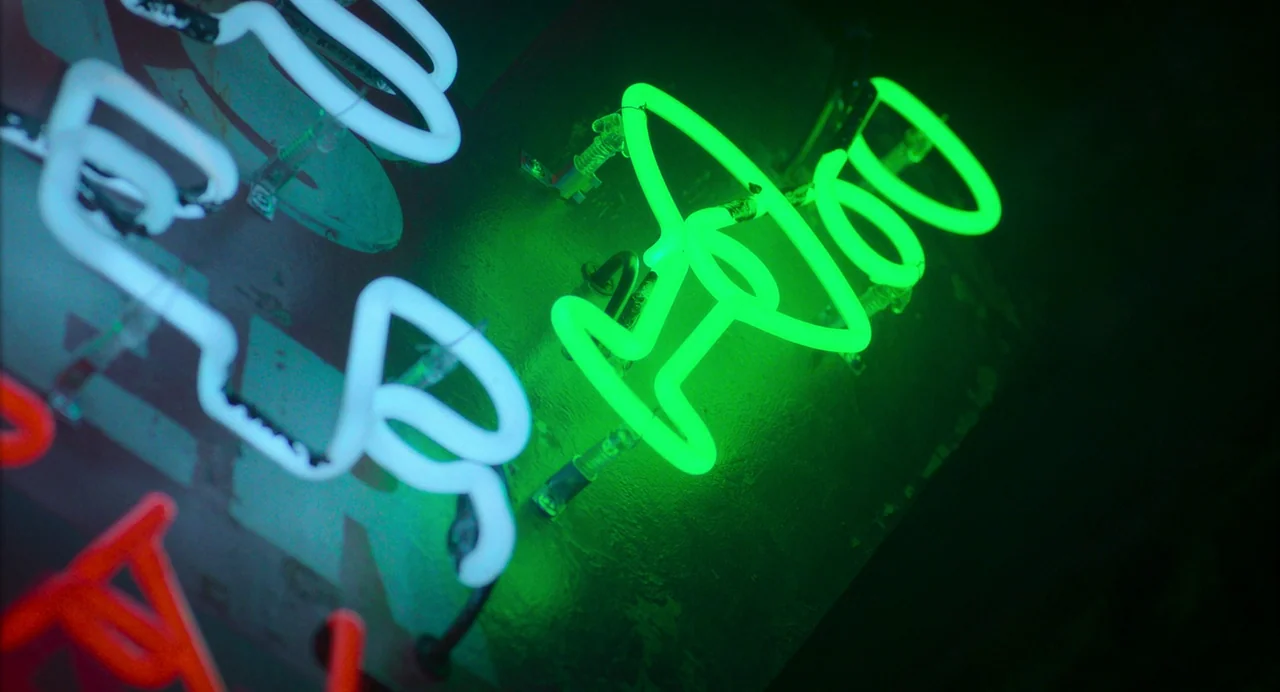 Drive-Away Dykes (2024) movie still: closeup — A close-up view shows glowing green and cyan neon tubes mounted on a dark surface. The glass tubes…