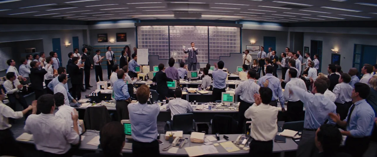 The Wolf of Wall Street (2013) movie still: wide — A crowded corporate brokerage office features rows of desks with old computer monitors. A man in…