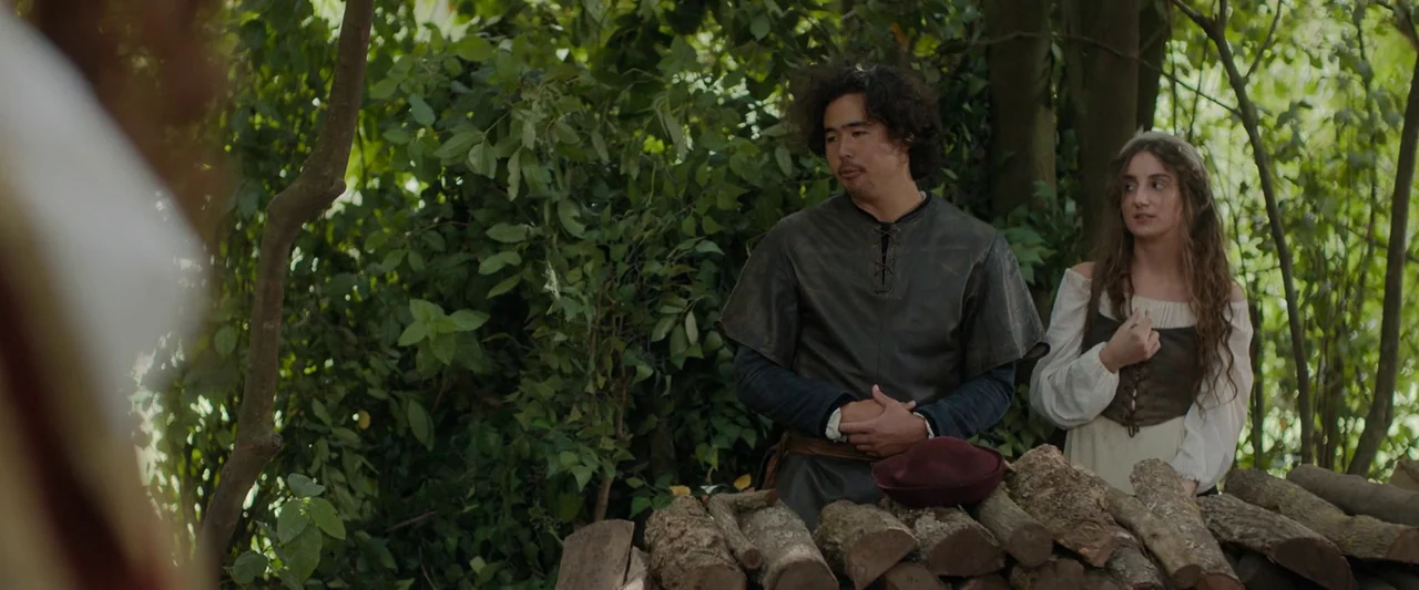 Rosaline (2022) movie still: medium — A man and woman stand in a dense, green wooded area behind stacked timber logs. The figures are dressed in…