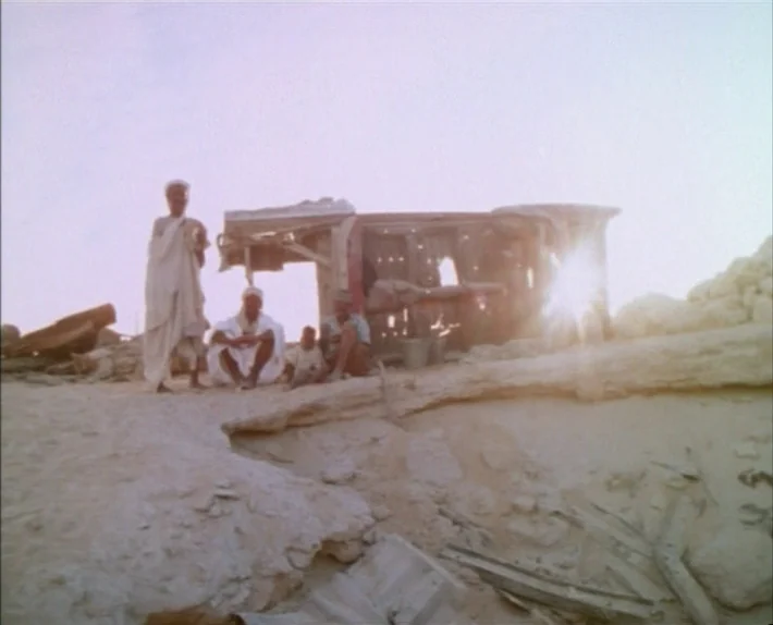 Fata Morgana (1971) movie still: wide — A remote desert landscape contains a dilapidated wooden structure made of salvaged materials. Three men sit…