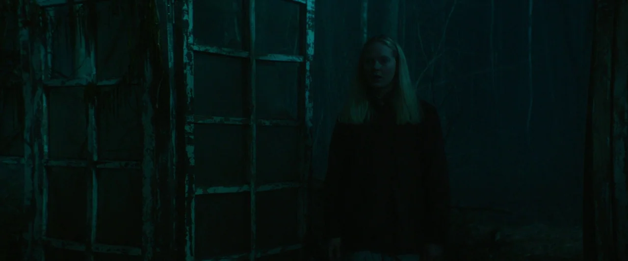 Vesper (2022) movie still: medium — A dark, wooded environment at night featuring an aged structure with grimy glass panes. A young woman with…