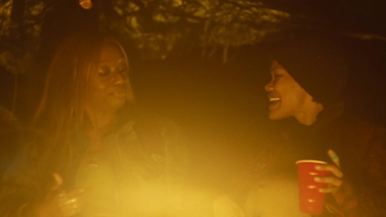 One Battle After Another (2025) movie still: medium — Two women socialize outdoors at night around a bright, glowing, off-camera bonfire. They smile…