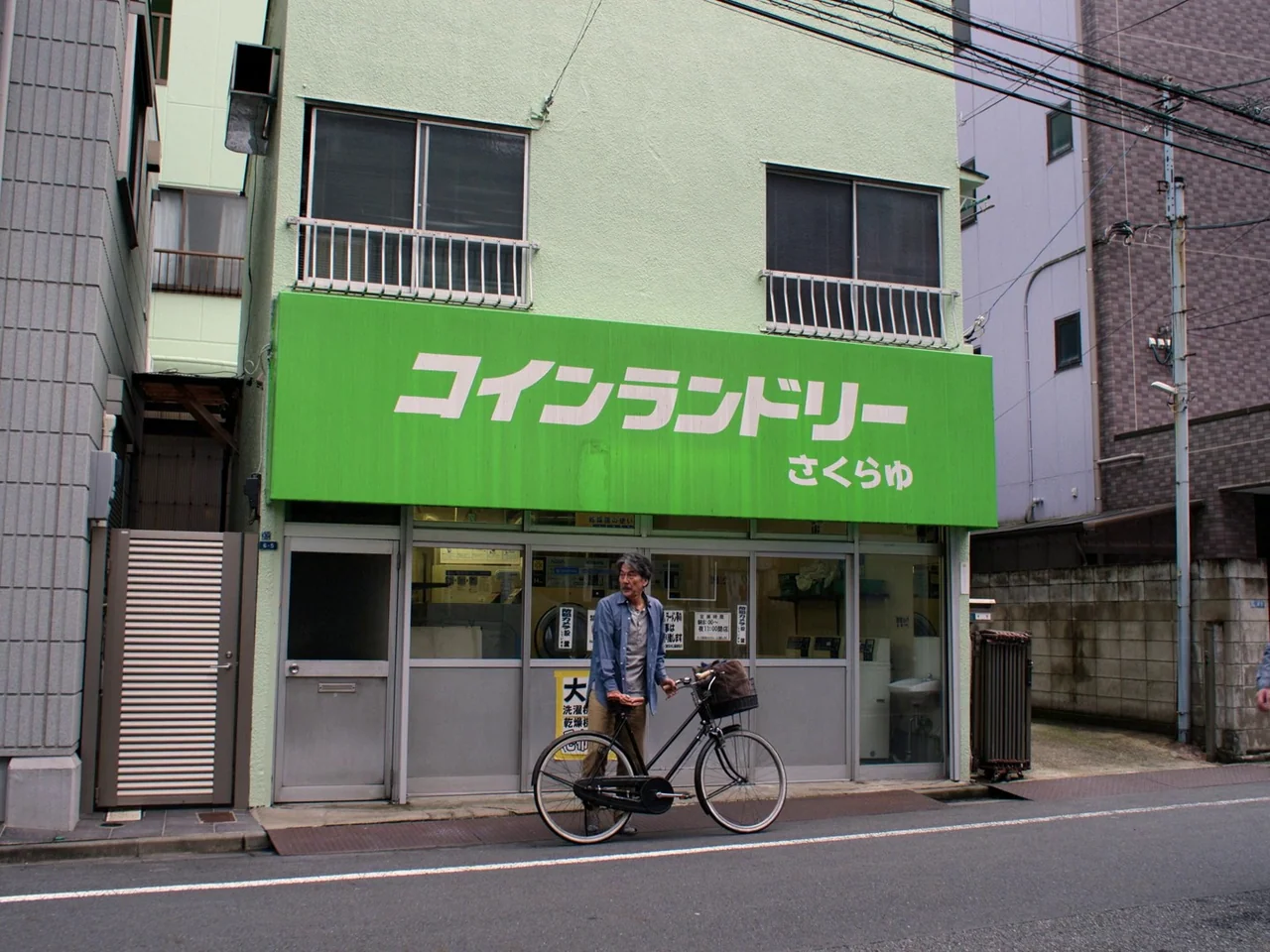 Perfect Days (2023) movie still: fullbody — A light green laundromat building stands along a quiet paved city street. A middle-aged man stands next…