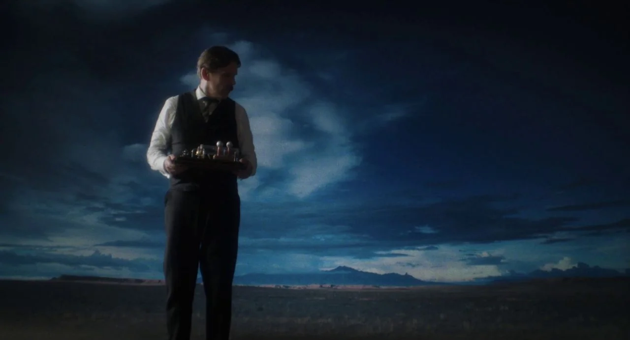 Tesla (2020) movie still: medium — A vast, dark desert landscape stretches under a moody, cloud-filled night sky. A man in period formal wear stands…