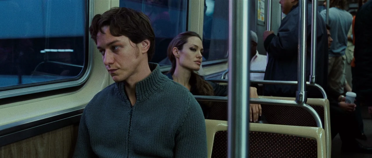 Wanted (2008) movie still: medium — A commuter train car at night features rows of patterned seats and metal railings. A young man sits in the…