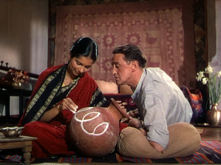 The River (1951) movie still: medium — An indoor space features a decorative quilt hanging on the wall and wooden floor mats. A man and a woman sit…