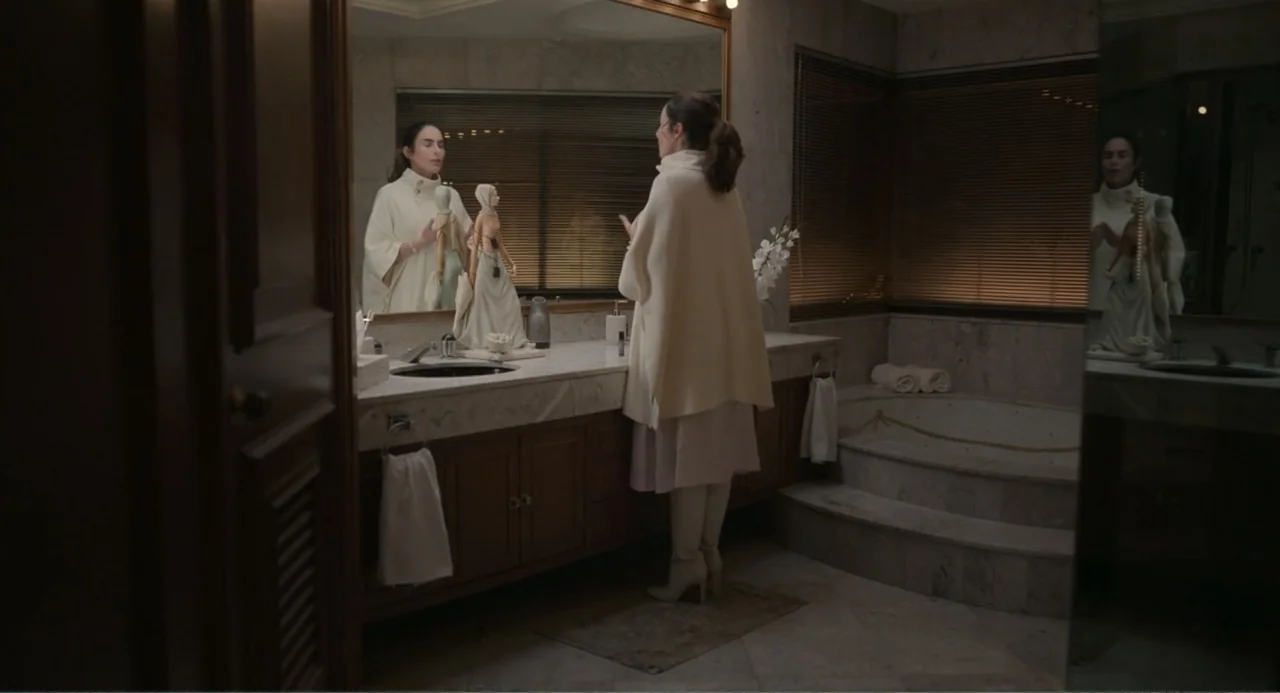 La posada (2023) movie still: medium — A marble-clad bathroom features a large vanity with an ornate figurine on the counter. A woman in a…