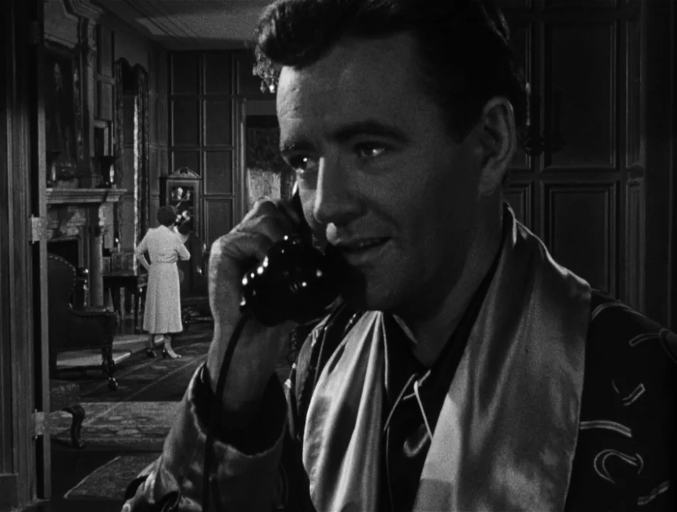 Strangers on a Train (1951) movie still: medium — A dark, wood-paneled room is lit with warm interior tones and deep shadows. A man in the…