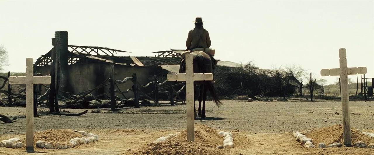 The Proposition (2005) movie still: wide — An arid, sun-scorched cemetery features three dirt graves marked by simple wooden crosses. A lone man…