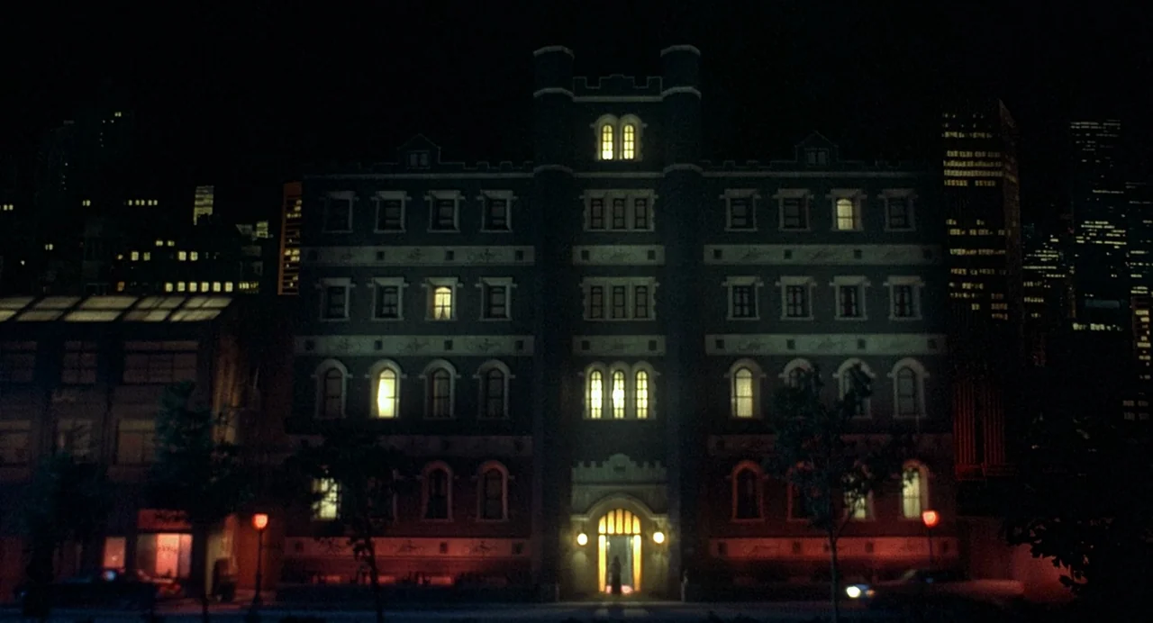 Inferno (1980) movie still: establishing — A dark brick building with arched windows stands against a distant city skyline. Multiple windows emit a…
