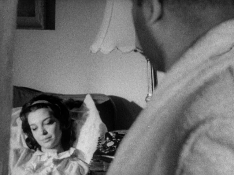 Shadows (1958) movie still: medium — A dimly lit domestic setting with a bed and a bedside lamp visible. A man stands in the foreground while a…