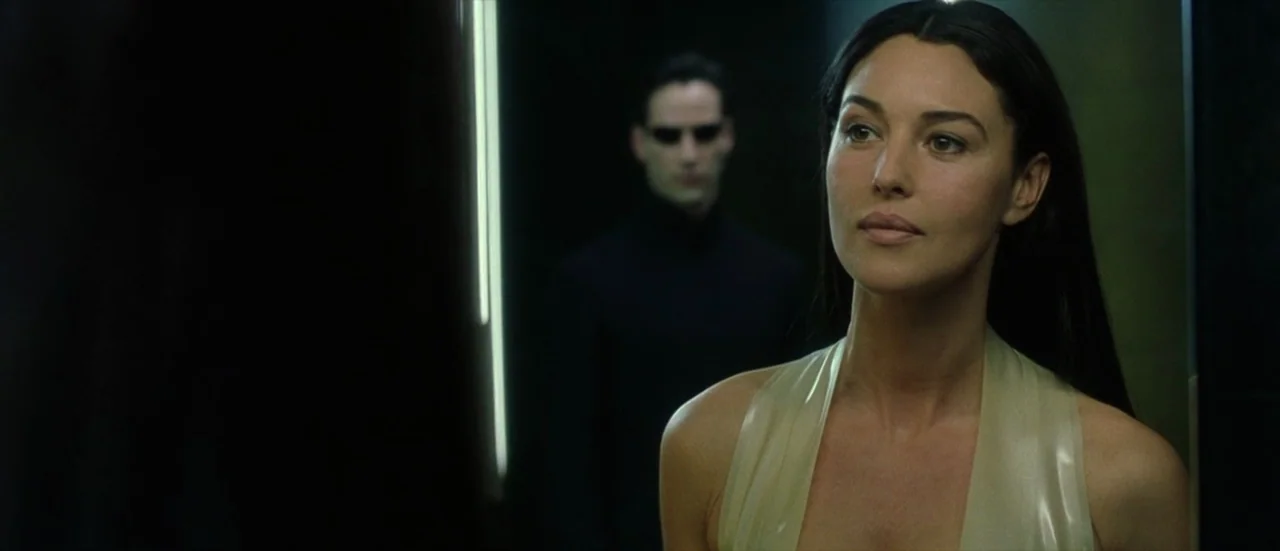 The Matrix Reloaded (2003) movie still: medium — A dark hallway with sleek black walls and vertical light strips in the background. A woman in a…