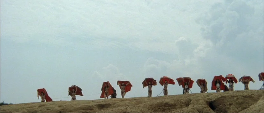 The Horrors of Malformed Men (1969) movie still: wide — A row of figures stands along the crest of a large, barren sand dune. They hold large red…