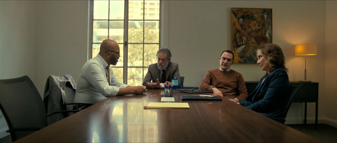 American Fiction (2023) movie still: wide — A professional conference room contains a long wooden table and modern office chairs. Four people sit…