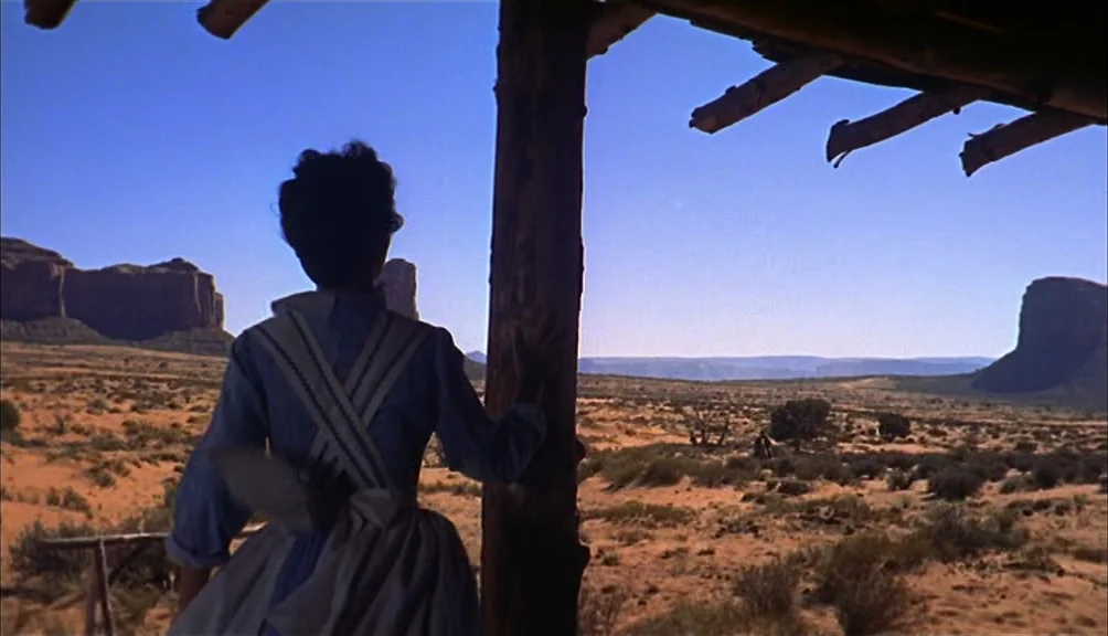 The Searchers movie still: medium — A wooden porch overlooks a vast, sun-drenched desert landscape with distant rock formations. A woman stands with…