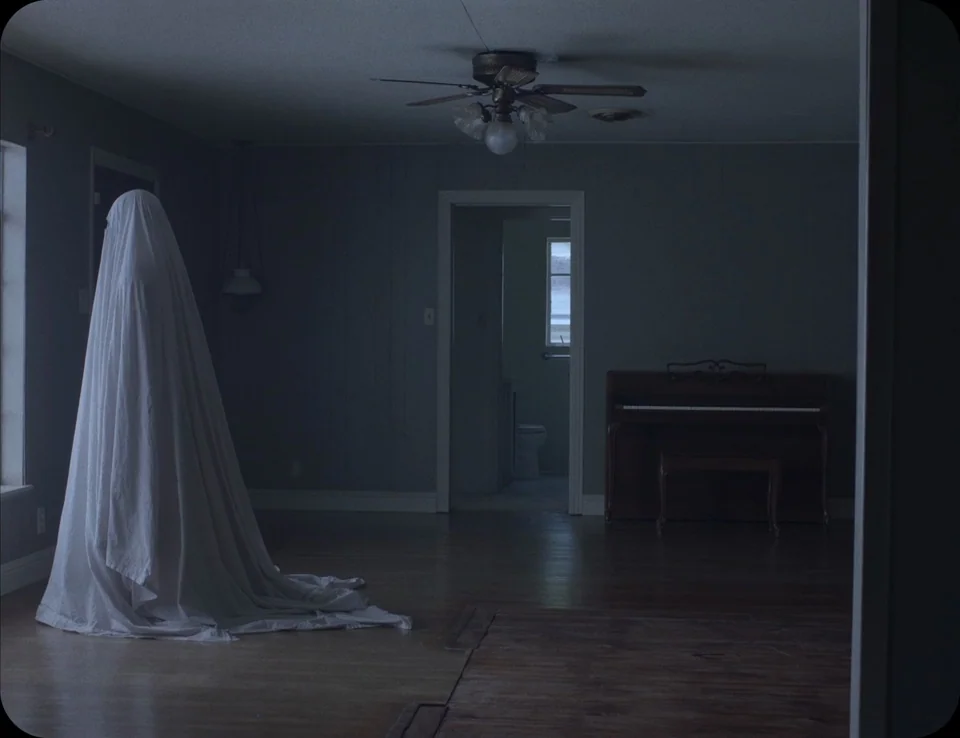 A Ghost Story (2017) movie still: fullbody — A quiet residential interior features a white sheeted figure standing near a side window. A piano rests…
