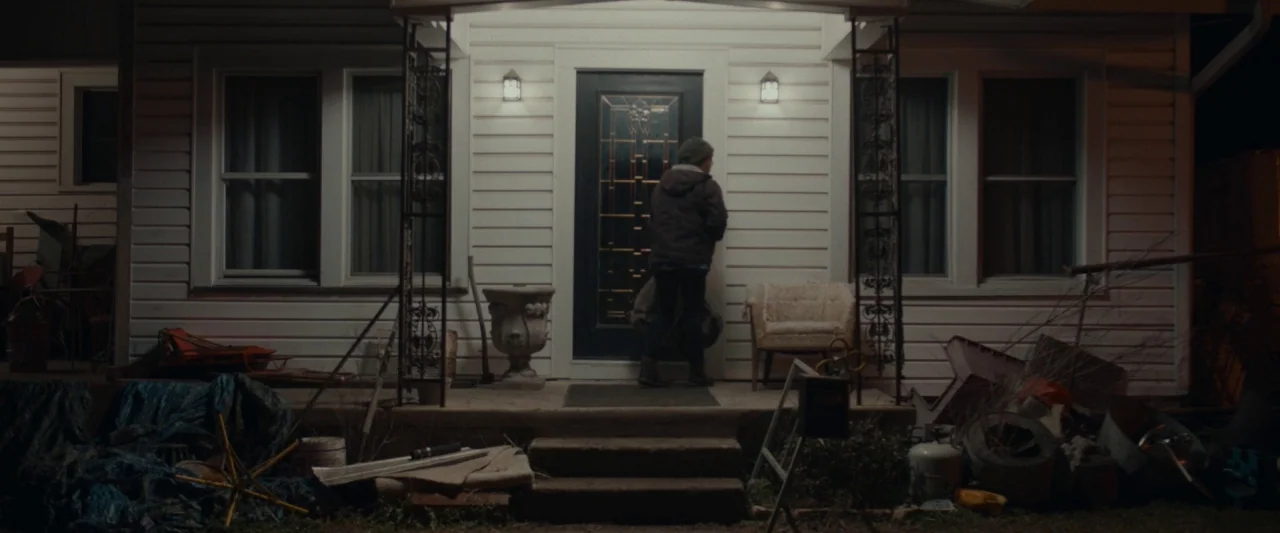 Little Woods (2018) movie still: fullbody — The front entrance of a suburban house is surrounded by assorted outdoor clutter. A person stands facing…