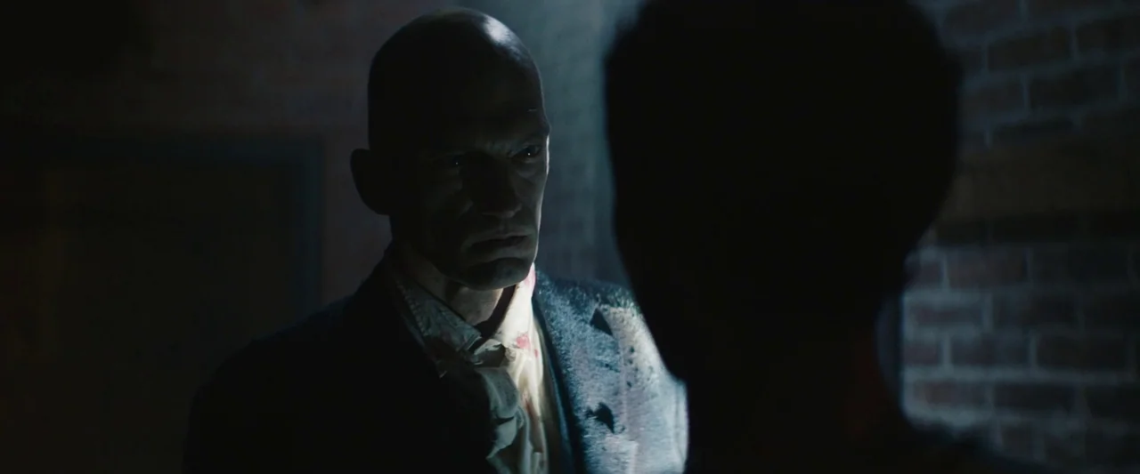 The Mad Hatter (2021) movie still: medium — A dimly lit industrial space features exposed brick walls and low ambient lighting conditions. A man…