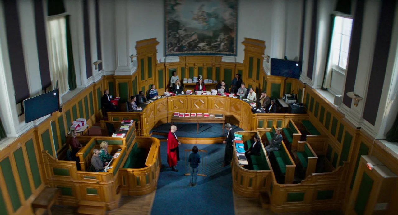 Anatomy of a Fall (2023) movie still: establishing — A spacious wood-paneled courtroom features tiered seating for participants and a large wall…