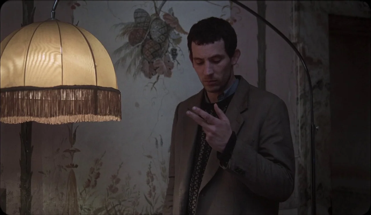 La Chimera (2023) movie still: medium — A dimly lit room features a wall with faded, hand-painted botanical wallpaper patterns. A man in a beige…