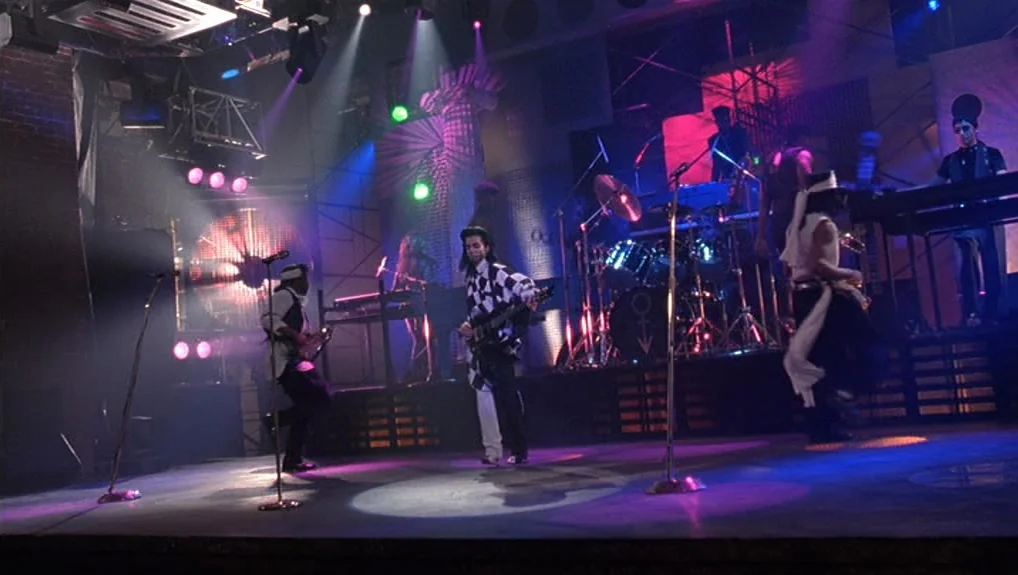 Graffiti Bridge (1990) movie still: wide — A dark concert stage features a musician in a checkered coat holding a guitar. The band members perform…