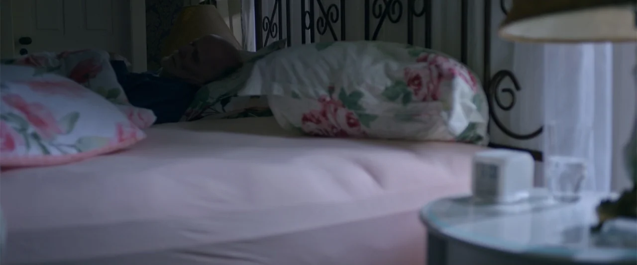 Dirty Little Secret (1998) movie still: medium — A bedroom contains a metal-framed bed with floral patterned pillows and pink sheets. An elderly man…