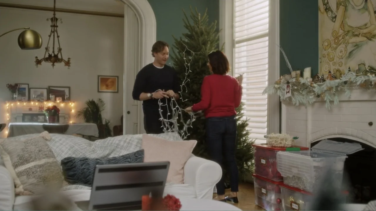 A Christmas Break (2020) movie still: medium — A living room features a fireplace, a white couch, and a large bare Christmas tree. A man and a woman…
