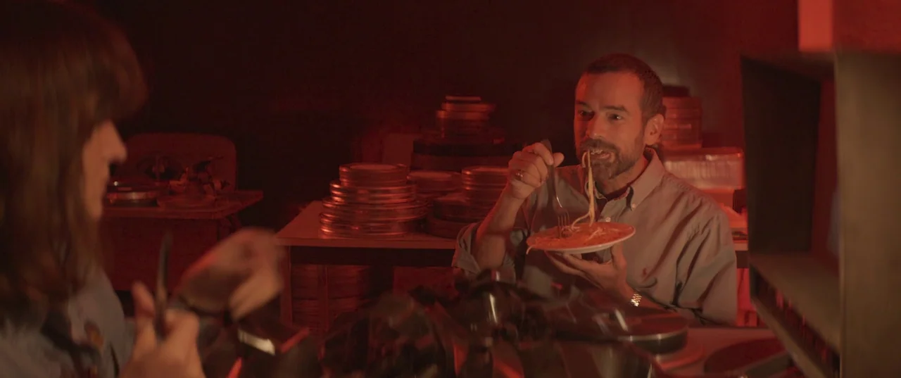 Daaaaaalí! (2023) movie still: medium — A dimly lit room filled with stacks of circular metal film canisters on tables. A man sits at a table eating…