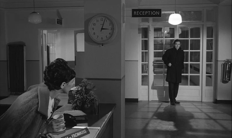 Lolita (1962) movie still: medium — An interior reception area features a clock on the wall and glass doors. A woman sits at a desk as a man enters…