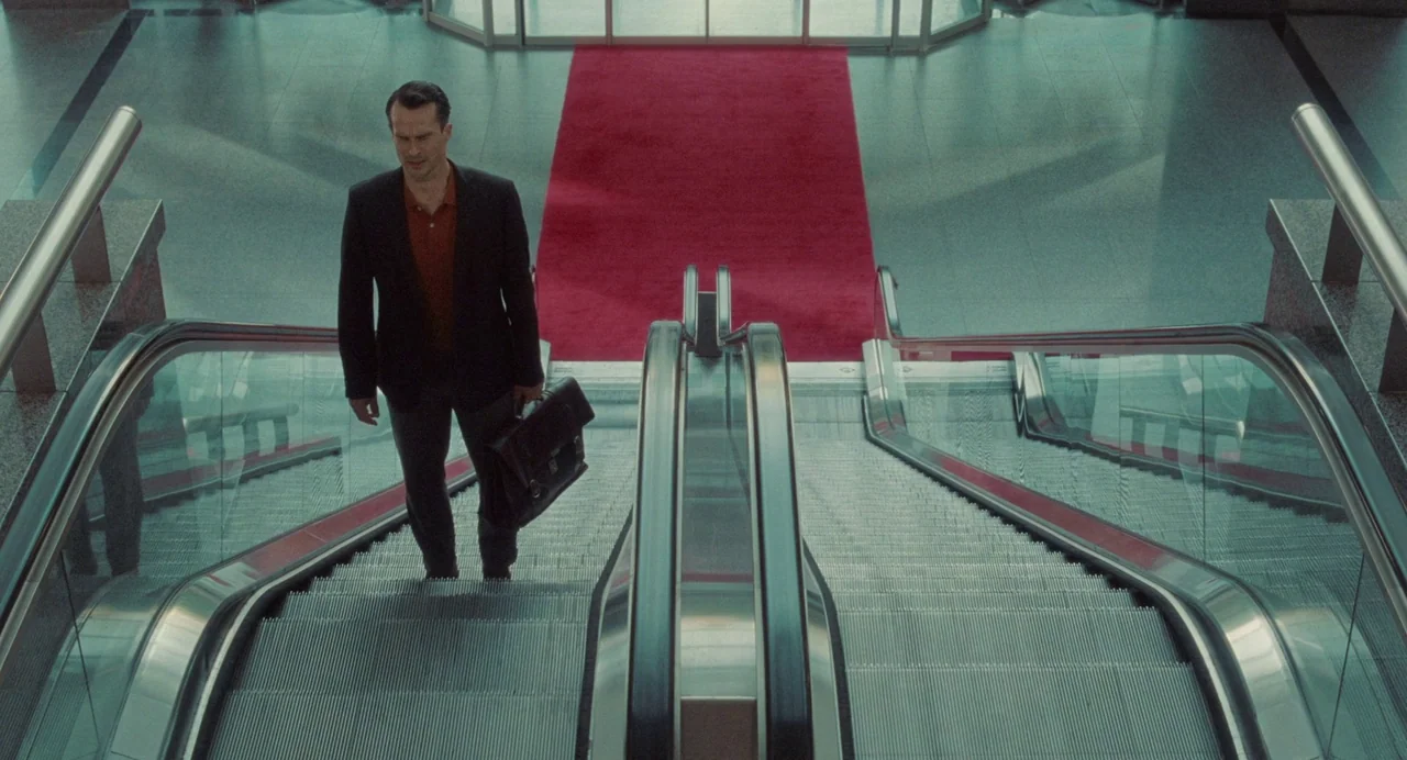 Babysitter (2022) movie still: fullbody — A man rides an escalator upward toward a floor with a red carpet. The modern architectural space features…