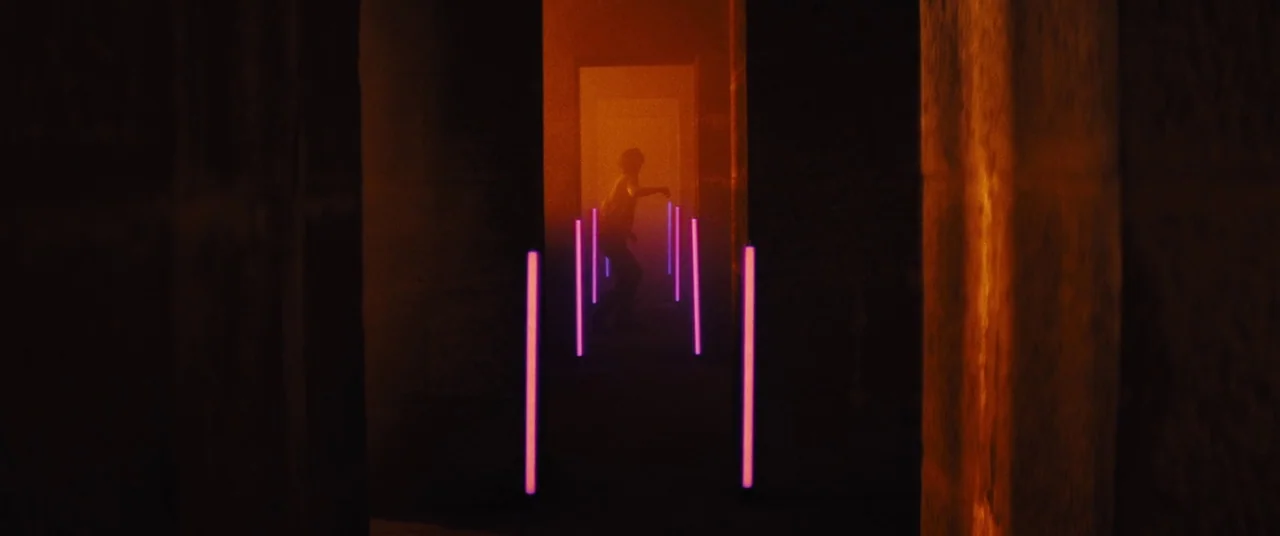 Daniel Isn’t Real movie still: wide — A dark, narrow corridor features stone-like walls illuminated by glowing purple neon light sticks. A person…